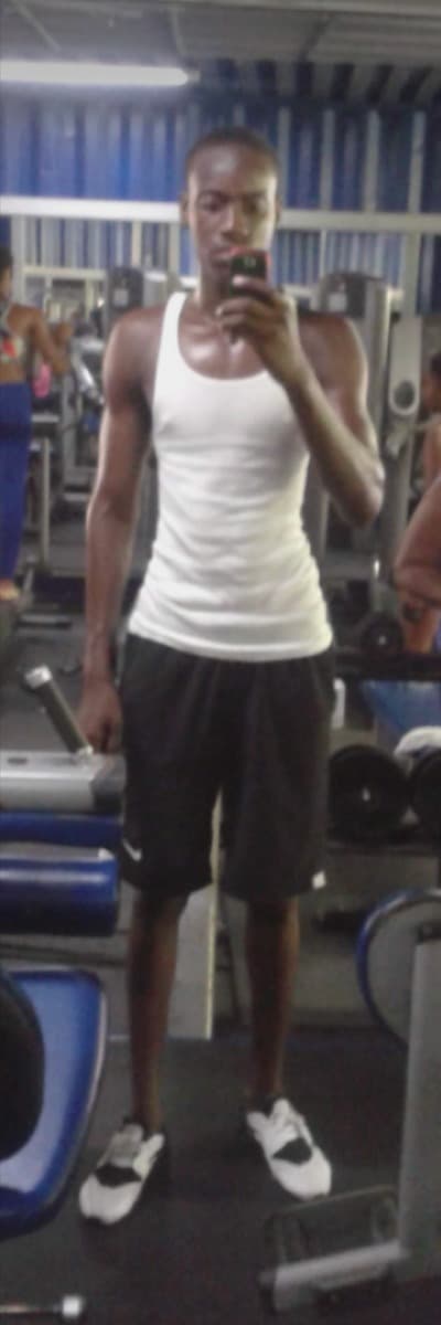 Nickoy Anthony in a gym mirror in October 2018, in a white tank top and shorts, holding a phone